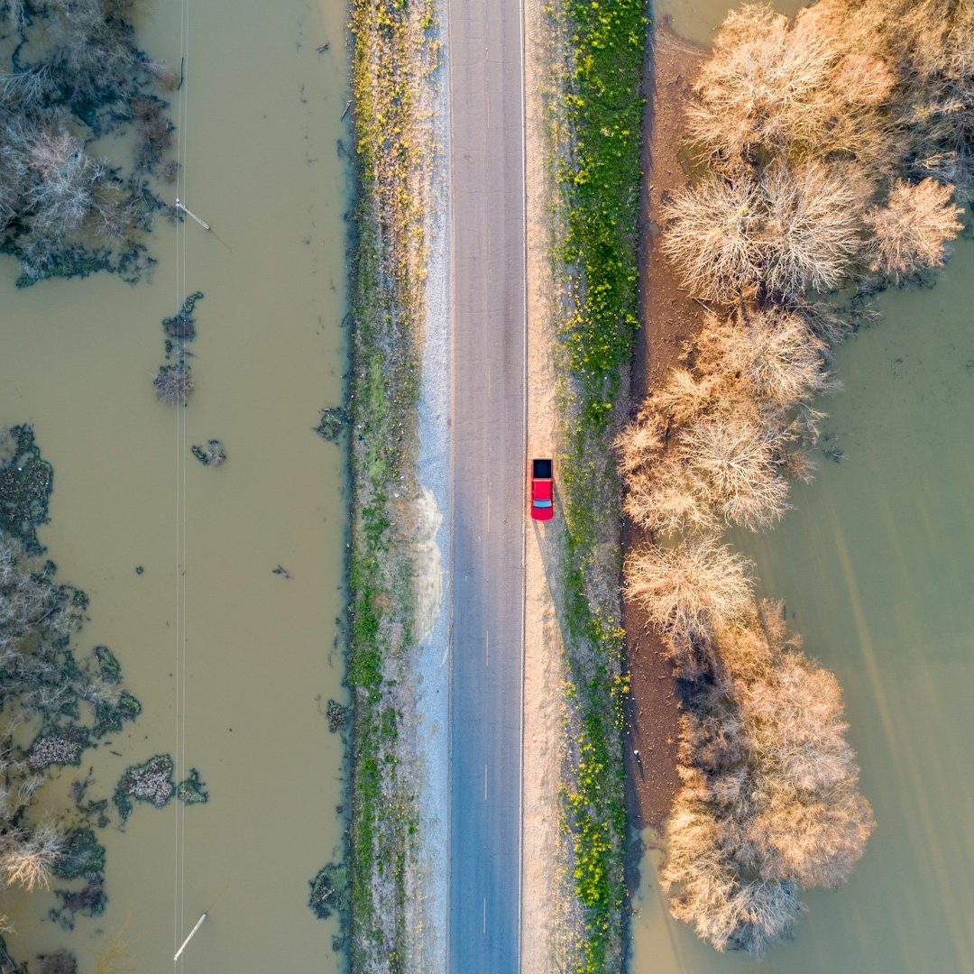 Rote Fahrzeug auf schmalem Weg zwischen Wasser und Bäumen.