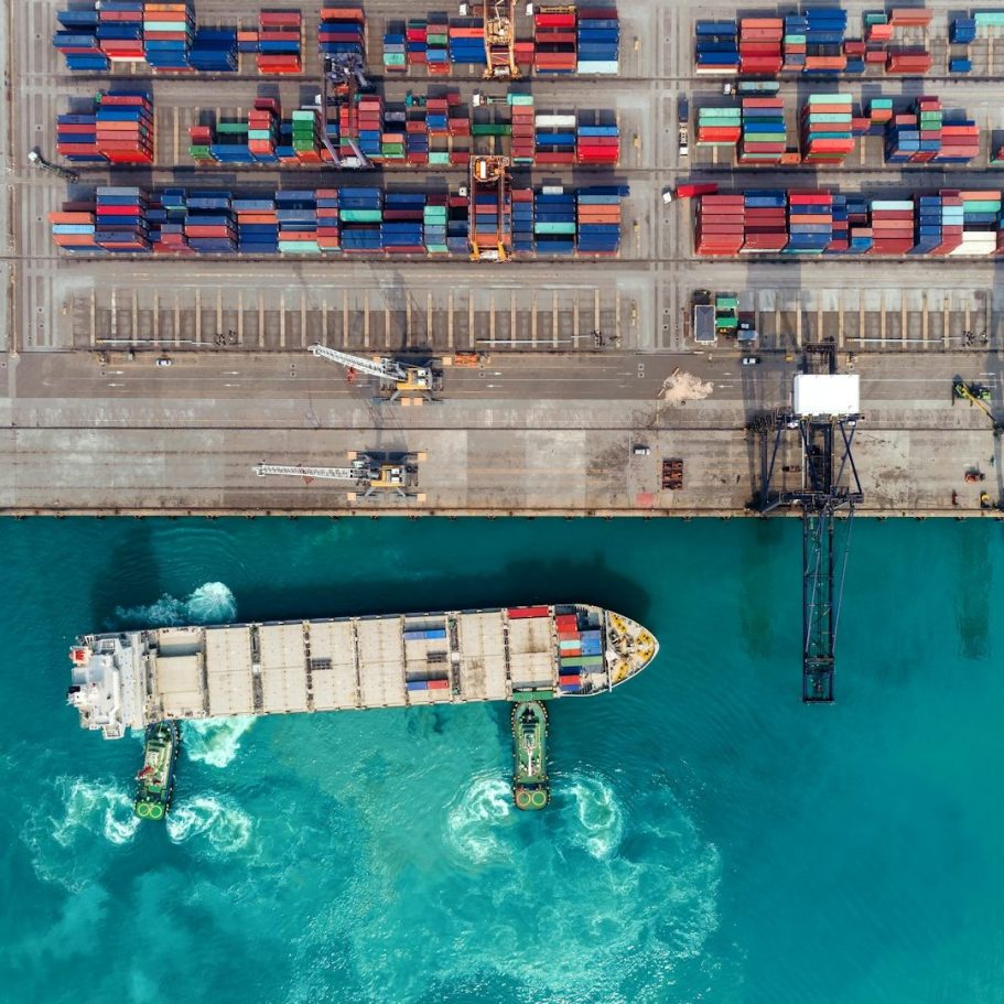 Containerfähre im Hafen, umgeben von Stapeln bunter Container und blauem Wasser.