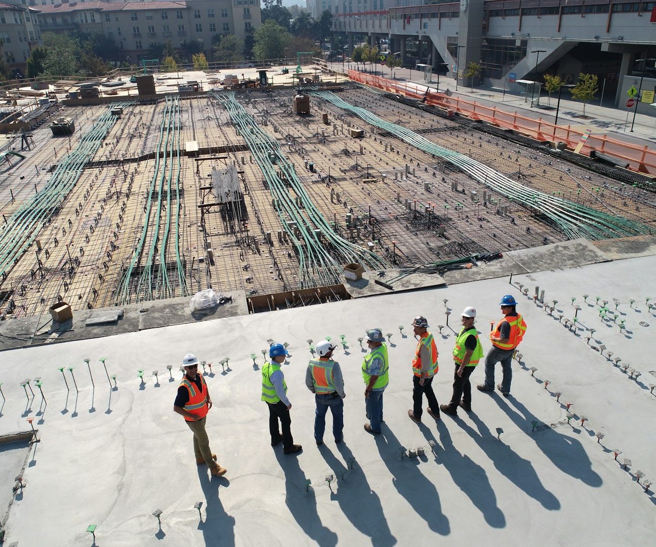 Sechs Bauarbeiter in Sicherheitswesten auf einer Baustelle mit Rohbau und grünen Rohren.